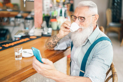 Bearded senior man drinking coffee while using smart phone in cafe