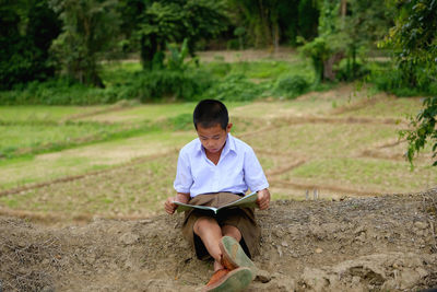 Full length of cute boy reading book while sitting on field against trees