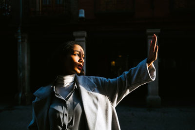 Full body of young ethnic female in stylish autumn outfit outstretching hand and covering face from sunlight while standing on pavement of old urban street