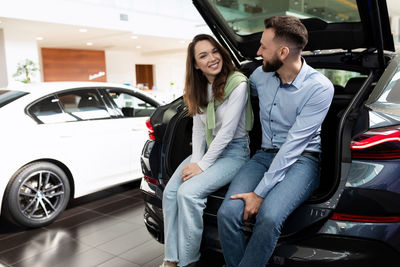 Happy couple sitting in car trunk at showroom