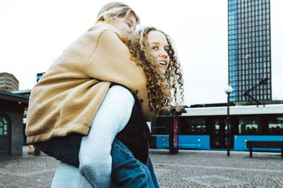 Girl carrying female friend piggyback on footpath while hanging out against clear sky