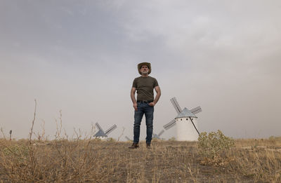 Full length of man with cowboy hat standing on field with spanish white windmills against sky