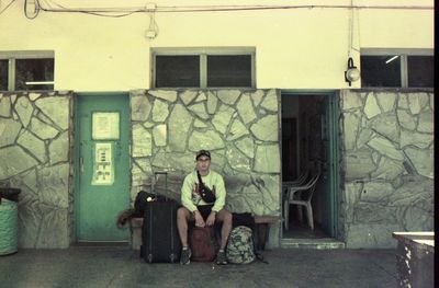 Portrait of man sitting with luggage on bench against wall