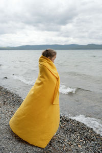 Side view of thoughtful girl standing by the sea wrapped in a yellow blanket on a cold day