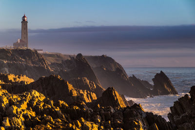 Lighthouse on rock formations by sea against sky during sunset