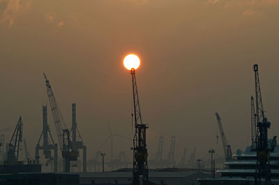 Cranes at harbor against sky during sunset