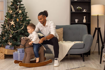 A young stylish father sits on a gray sofa, rolls his little son on a brown rocking horse, at home