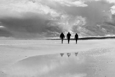 Silhouette friends standing at sandy beach against cloudy sky