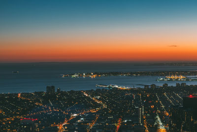 Aerial view of illuminated city by sea against sky at dusk