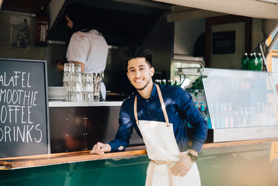 Portrait of smiling owner standing by food truck while female coworker working in background