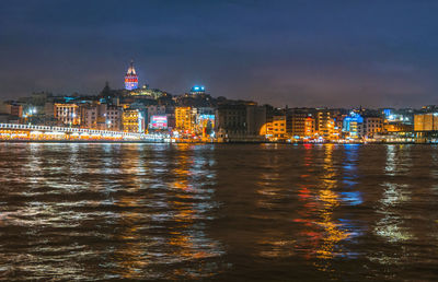 Illuminated buildings by river against sky at night