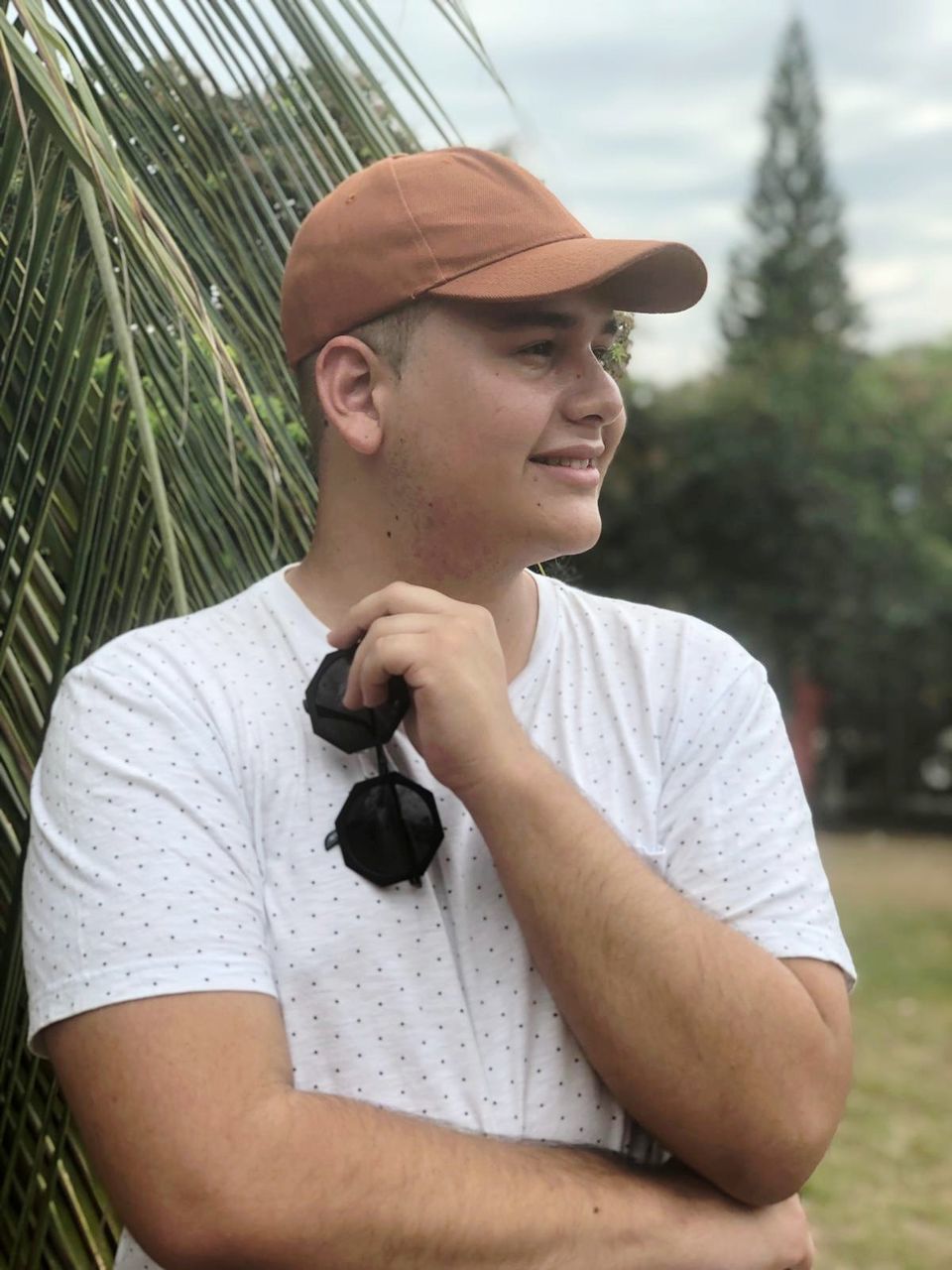 one person, real people, leisure activity, waist up, front view, young men, plant, casual clothing, lifestyles, young adult, hat, portrait, men, smiling, males, day, looking, tree, focus on foreground, outdoors, contemplation, mature men, teenager, adolescence