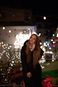 Portrait of smiling young woman standing against illuminated wall at night