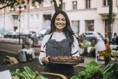 Portrait of smiling vendor holding vegetable tray while standing at farmer's market