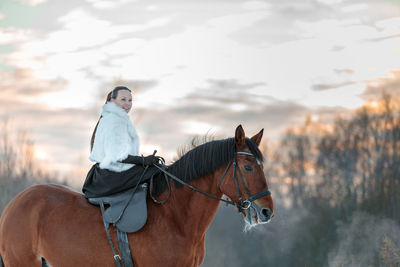 A girl in a white cloak rides a brown horse in winter. golden hour, setting sun.