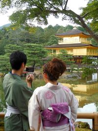 Rear view of man and woman photographing by pond against building