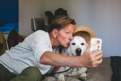 Senior man kissing dog while taking selfie on smart phone in bedroom at home