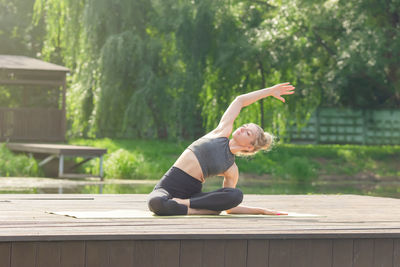 A woman sitting on a wooden platform by a pond in summer and performing a body tilt sideways