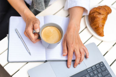 A woman works on a laptop, takes notes, and drinks coffee during an online morning meeting at a cafe