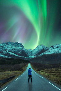 Distant view of explorer standing on asphalt road in snowy mountains under dark night sky with bright green polar lights in norway