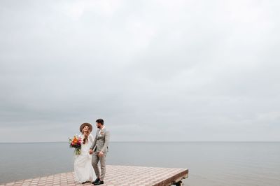 Newly married couple walking on pier over lake against cloudy sky