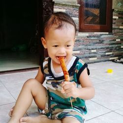 Cute baby boy eating sausage while sitting on floor against house