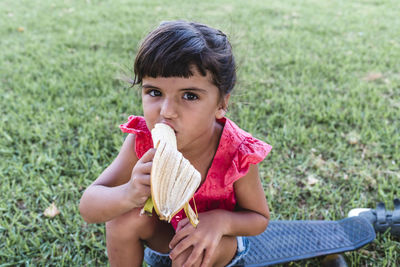 Little girl eating banana sitting outdoors on the grass in a park.