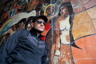 Young man wearing sunglasses standing against graffiti wall