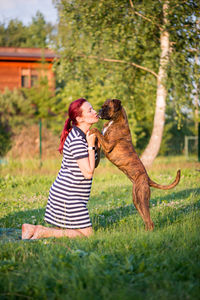 Mid adult woman playing with dog while kneeling on grassy field against sky at park