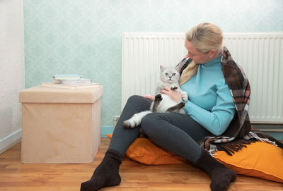 Woman sitting with cat under blanket warming near an electric heater,