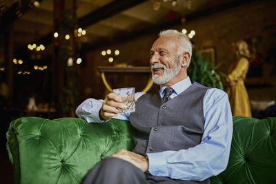 Portrait of elegant senior man sitting on couch in a bar holding tumbler