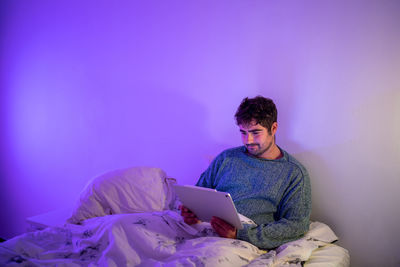 Man using digital tablet sitting on bed against wall at home