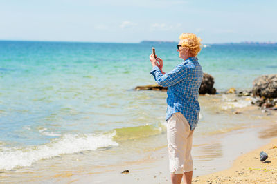 Elderly woman standing on the beach and taking photo on a mobile phone