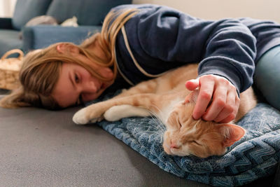 A young adolescent girl lying on a couch finds comfort by snuggling and petting her tabby cat