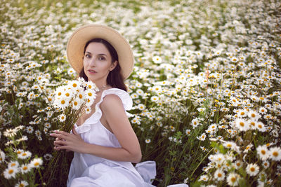 Beautiful young brunette woman in a hat and a white dress standing on a chamomile field at sunset