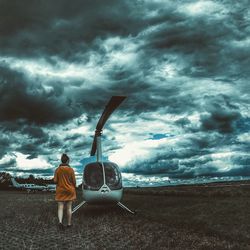 Rear view of woman standing by helicopter on field against cloudy sky