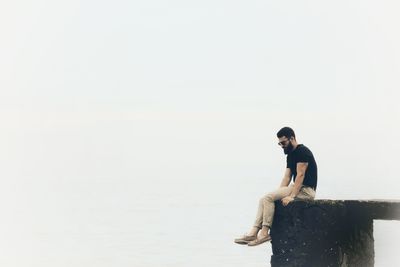 Full length of man sitting on pier at beach against clear sky