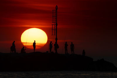 Silhouette people on rocks by sea against orange sky