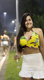 Portrait of smiling young woman holding champagne flute while standing outdoors at night