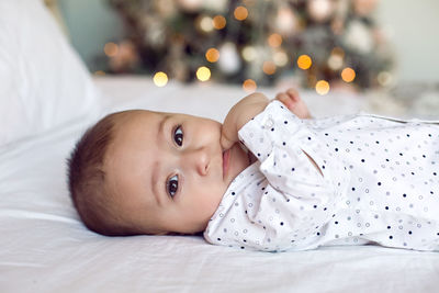 Baby boy six months lying on the bed in a white shirt next to the christmas tree