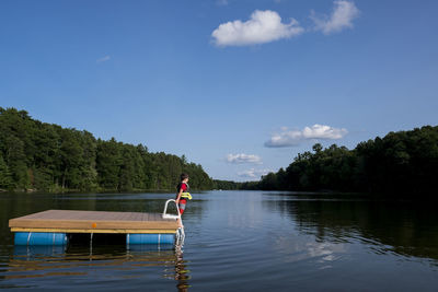 Side view of boy standing on floating platform in lake at forest against sky