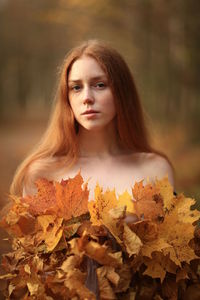 Portrait of young woman covered with leaves standing at forest during autumn