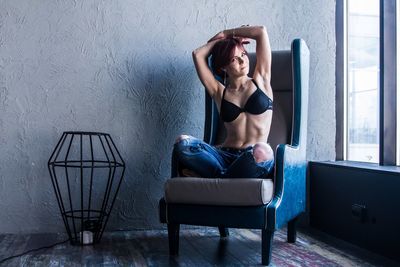 Portrait of woman wearing bra while sitting on armchair by window at home