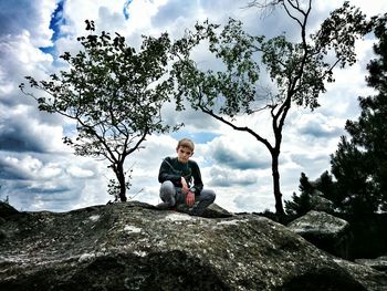 Low angle portrait of boy crouching on rock against cloudy sky