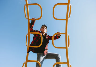 Low angle view of boy climbing on metallic steps against clear blue sky at playground during sunny day