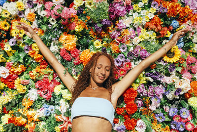 Smiling young woman with arms raised standing in front of flower wall on sunny day