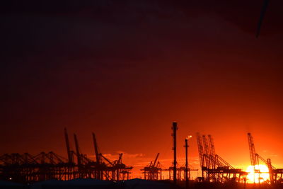 Silhouette cranes at harbor against orange sky