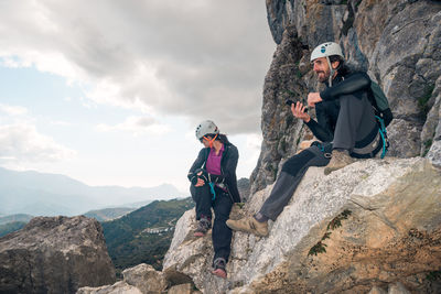 Concept: adventure. pair of climbers with helmet and harness. resting sitting on a rock. using the smart phone while looking at the horizon. via ferrata in the mountains.