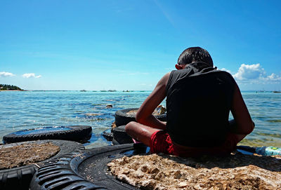 Rear view of boy sitting at beach by tiers against blue sky