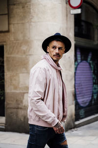 Stylish man wearing hat looking away while walking on street against building